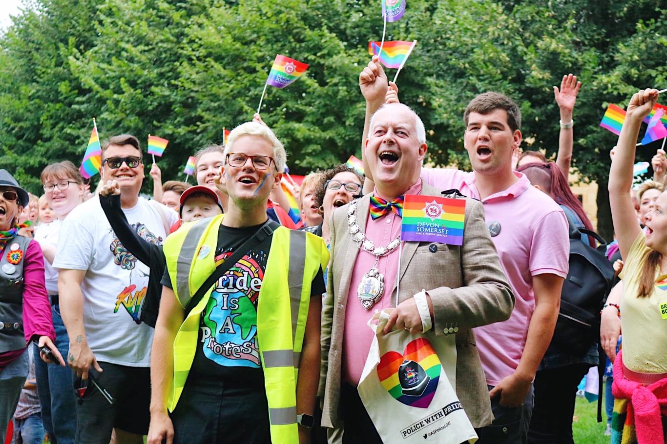 Mobilising and energising the LGBTQIA+ community in Yeovil. JK stands next to the Mayor of Yeovil who is wearing ceremonial chains and carrying a 'pride' flag, in front of a larger crowd cheering.