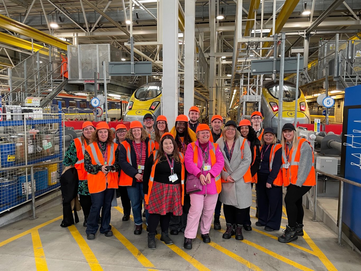 Creating experiences that have an impact, JK is a self-starter who organises. JK stands with a group of colleagues, all in orange hi-vis inside Eurostar's depot in East London.